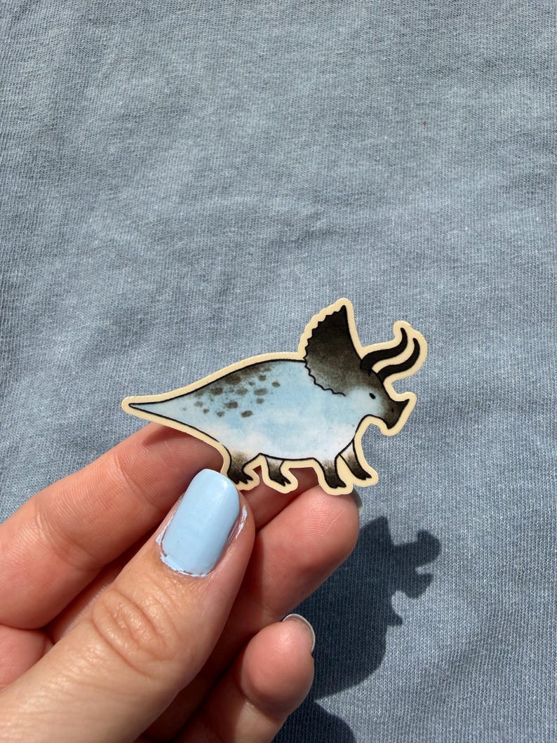 May include: A sticker of a triceratops dinosaur. The dinosaur is light blue with dark blue spots and a dark gray frill and horns. The sticker has a light brown border and is on a light blue background.