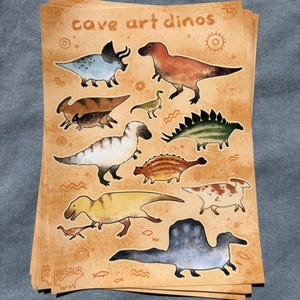 May include: A stack of stickers featuring various dinosaurs in a cave art style. The stickers have a tan background with the words "cave art dinos" and "Anisaur" printed on them. The dinosaurs are in various colors.
