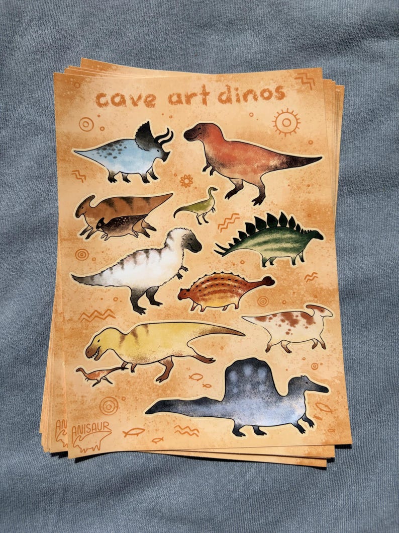 May include: A stack of stickers featuring various dinosaurs in a cave art style. The stickers have a tan background with the words "cave art dinos" and "Anisaur" printed on them. The dinosaurs are in various colors.