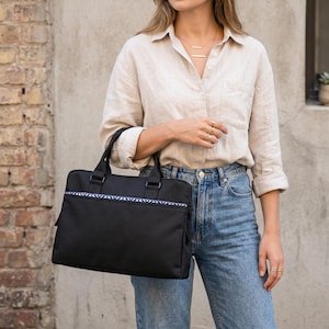 May include: A black briefcase-style handbag with a blue and white patterned accent along the top. The bag has two handles and a rectangular shape. The person in the image is wearing a light-colored button-down shirt and blue jeans.