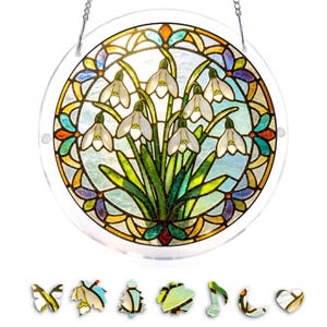 May include: A round, stained glass window featuring a cluster of white snowdrop flowers with green leaves. The window has a colorful border with blue, yellow, and orange accents. A silver chain is attached for hanging.
