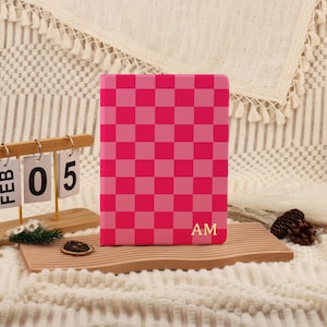 May include: A pink and light pink checkered pattern notebook with gold-colored initials "AM" on the bottom right corner. The notebook is displayed on a wooden tray with a calendar showing "FEB 05".