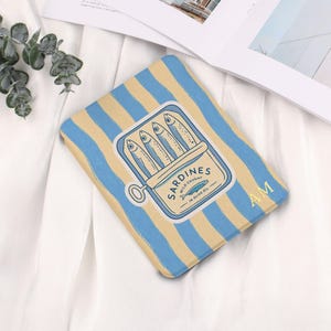 Puede incluir: Funda rectangular con un diseño de rayas azules y beige. Presenta una ilustración de una lata de sardinas con el texto "Sardines Wild Caught in Olive Oil". Las iniciales "AM" están en la esquina inferior derecha.