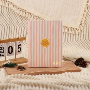 Puede incluir: Un diario a rayas rosas y blancas con una ilustración de un sol sonriente y las iniciales "AM". El diario es rectangular y tiene una cubierta blanda. Un calendario de madera muestra "FEB 05" en el fondo.