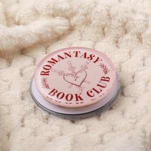 May include: A round, pink button with the text "ROMANTASY BOOK CLUB" in maroon. The button features a heart pierced by two swords. The button is set on a silver-colored base, resting on a white, textured surface.