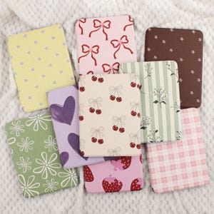 Può includere: Una collezione di quaderni rettangolari con vari motivi. I disegni includono fiocchi rosa, ciliegie, fragole, cuori, fiori e quadretti vichy. I colori includono rosa, giallo, verde, viola e marrone. I quaderni sono disposti su una superficie bianca strutturata.