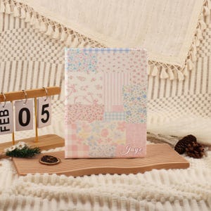 Può includere: Un diario patchwork floreale con una combinazione di colori rosa tenue, blu e bianco. La copertina presenta una varietà di motivi, tra cui righe, quadretti e stampe floreali. Il nome "Jaye" è scritto in corsivo in basso a destra.
