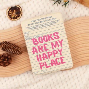 May include: A notebook with the text "BOOKS ARE MY HAPPY PLACE" in pink letters. The notebook has a vintage library card design with lines and text. The background includes a wooden surface, pine cones, and a dried orange slice.