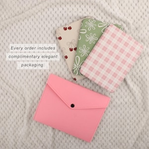 Puede incluir: Un estuche rosa tipo sobre con cierre de bot&oacute;n a presi&oacute;n negro, junto a tres cuadernos estampados. Los cuadernos presentan dise&ntilde;os de cerezas, flores y cuadros de vichy. El texto dice: Cada pedido incluye un embalaje elegante de cortes&iacute;a.