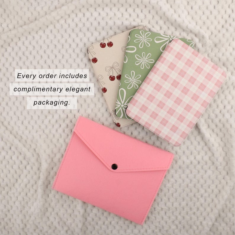 Puede incluir: Un estuche rosa tipo sobre con cierre de bot&oacute;n a presi&oacute;n negro, junto a tres cuadernos estampados. Los cuadernos presentan dise&ntilde;os de cerezas, flores y cuadros de vichy. El texto dice: Cada pedido incluye un embalaje elegante de cortes&iacute;a.