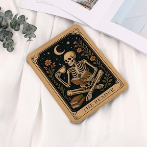 May include: A rectangular tarot card with a skeleton reading a book and holding a cup, under a crescent moon and stars. The card has a brown border and the words "THE READER" at the bottom.