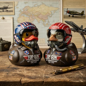 May include: Two rubber duck figurines dressed as pilots, one in a blue helmet labeled "Maverick" and the other in a red helmet labeled "Goose." Both wear aviator jackets with "Top Gun" and "40th Anniversary 1986-2026" printed on them. A vintage pen rests on the wooden surface.