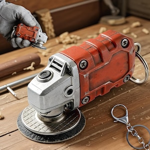 Può includere: Un portachiavi smerigliatrice angolare rosso e argento e una smerigliatrice angolare più grande su una superficie di legno. Il portachiavi è tenuto in mano da un guanto. La smerigliatrice più grande ha un disco nero e una testa in metallo argentato.