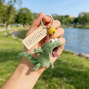 May include: A green plush lizard keychain with a wide open mouth and pink tongue. The keychain includes a small yellow bell and a wooden tag that reads "zoomy". The background shows a lake and trees.