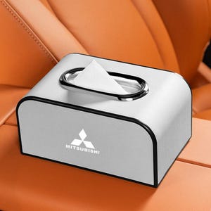 K&ouml;nnte beinhalten: Eine silberne Taschentuchbox mit schwarzem Rand und einer verchromten &Ouml;ffnung. Das Mitsubishi-Logo ist auf der Vorderseite abgebildet. Die Box steht auf einer beigen Oberfl&auml;che, wahrscheinlich in einem Fahrzeug.