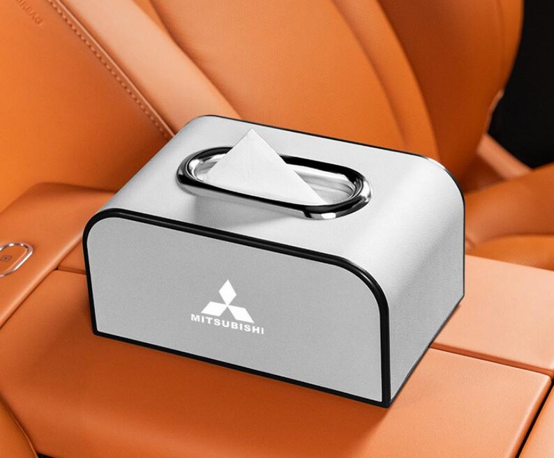 K&ouml;nnte beinhalten: Eine silberne Taschentuchbox mit schwarzem Rand und einer verchromten &Ouml;ffnung. Das Mitsubishi-Logo ist auf der Vorderseite abgebildet. Die Box steht auf einer beigen Oberfl&auml;che, wahrscheinlich in einem Fahrzeug.