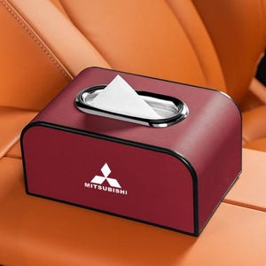K&ouml;nnte beinhalten: Eine weinrote Taschentuchbox mit einer silbernen ovalen &Ouml;ffnung und dem Mitsubishi-Logo. Die Box ist rechteckig mit abgerundeten Kanten und befindet sich auf der Mittelkonsole eines Autos. Wei&szlig;e Taschent&uuml;cher sind sichtbar.