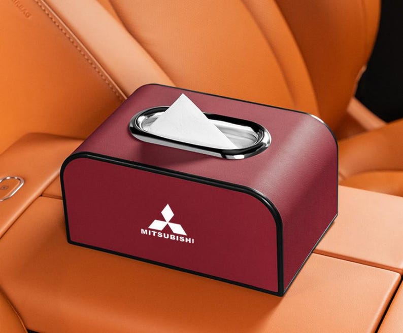 K&ouml;nnte beinhalten: Eine weinrote Taschentuchbox mit einer silbernen ovalen &Ouml;ffnung und dem Mitsubishi-Logo. Die Box ist rechteckig mit abgerundeten Kanten und befindet sich auf der Mittelkonsole eines Autos. Wei&szlig;e Taschent&uuml;cher sind sichtbar.