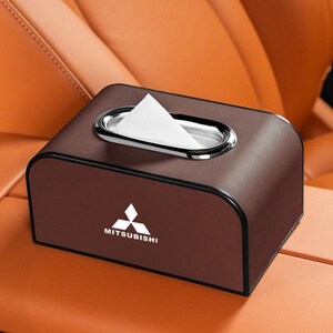 K&ouml;nnte beinhalten: Eine braune Tissuebox aus Kunstleder mit Chromrand um die &Ouml;ffnung. Das Mitsubishi-Logo ist auf der Vorderseite abgebildet. Die Box ist rechteckig mit abgerundeten Kanten.