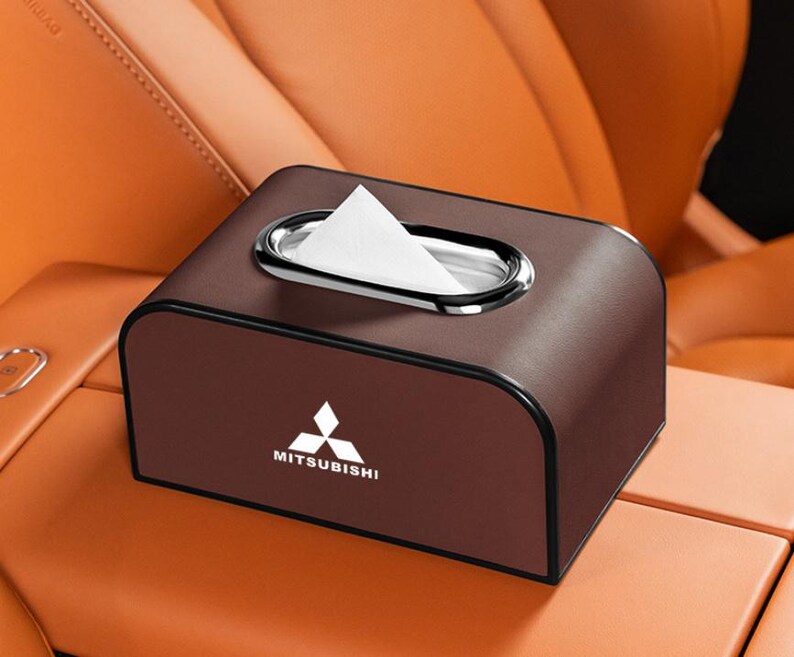 K&ouml;nnte beinhalten: Eine braune Tissuebox aus Kunstleder mit Chromrand um die &Ouml;ffnung. Das Mitsubishi-Logo ist auf der Vorderseite abgebildet. Die Box ist rechteckig mit abgerundeten Kanten.