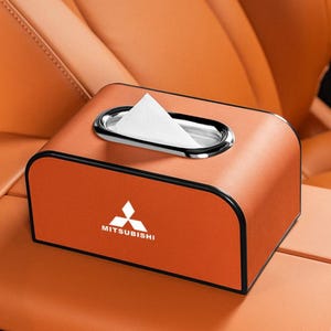 K&ouml;nnte beinhalten: Orangefarbene Taschentuchbox mit silberner Zierleiste um die &Ouml;ffnung. Die Box hat das Mitsubishi-Logo und den Text auf der Vorderseite. Die Box befindet sich auf der Mittelkonsole eines Autos.