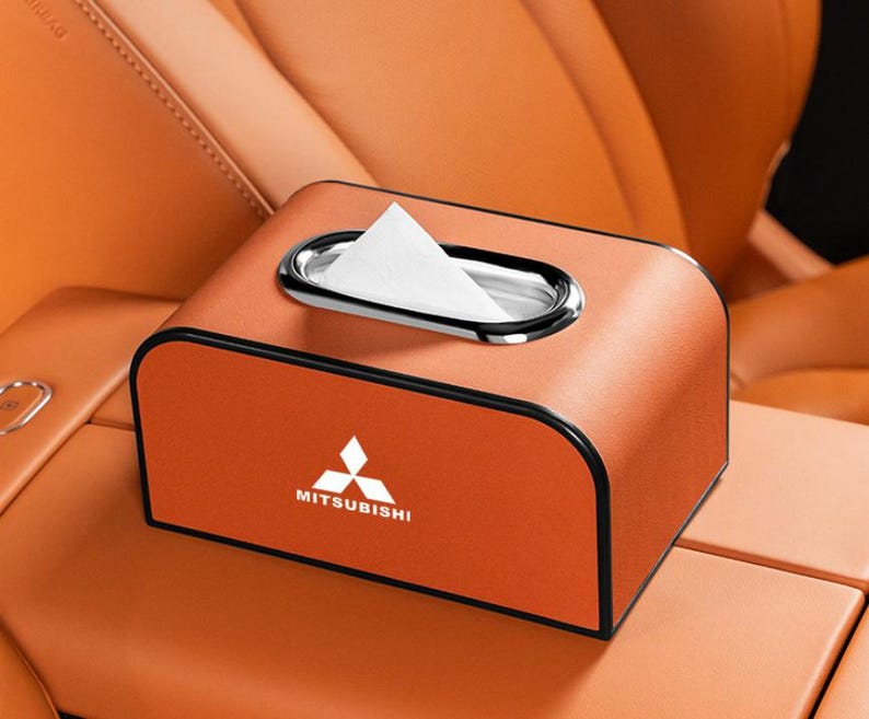 K&ouml;nnte beinhalten: Orangefarbene Taschentuchbox mit silberner Zierleiste um die &Ouml;ffnung. Die Box hat das Mitsubishi-Logo und den Text auf der Vorderseite. Die Box befindet sich auf der Mittelkonsole eines Autos.