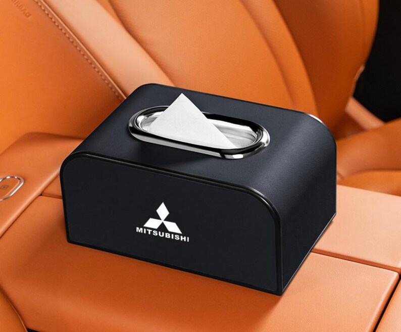 K&ouml;nnte beinhalten: Schwarze Taschentuchbox mit silberner Zierleiste und Mitsubishi-Logo. Wei&szlig;e Taschent&uuml;cher sind sichtbar. Die Box steht auf einer braunen Oberfl&auml;che.