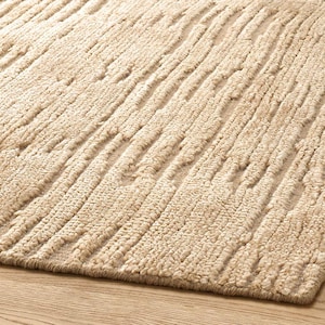 Puede incluir: Primer plano de una alfombra beige texturizada con un diseño tejido. La alfombra presenta líneas verticales en relieve en su superficie, creando un patrón táctil y visualmente interesante. El color neutro y la textura de la alfombra la hacen adecuada para varios estilos de diseño de interiores.