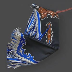 Puede incluir: Chaps de cuero negro con intrincados detalles en marrón y plata. Los chaps presentan flecos azules y plateados a lo largo de los bordes y un cinturón de cuero marrón. El diseño incluye recortes y costuras decorativas.