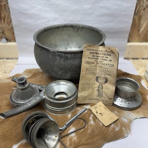 Puede incluir: Un surtido de piezas de separador de crema antiguas, que incluyen un cuenco de metal grande, varios embudos y un colador. La imagen también muestra un manual de instrucciones titulado "Assembling and Operating Instructions and Parts List for Economy Chief, Jr. Cream Separator."
