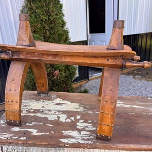 May include: A wooden stool with a curved seat and four arched legs, featuring metal studs along the base and sides. The structure includes two vertical wooden posts extending upward from the seat, each topped with a metal cap and secured by bands.