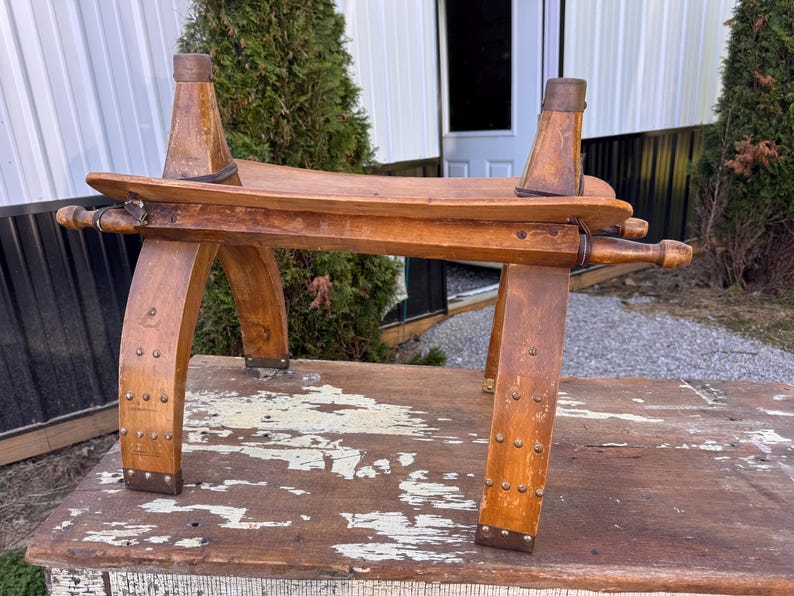 May include: A wooden stool with a curved seat and four arched legs, featuring metal studs along the base and sides. The structure includes two vertical wooden posts extending upward from the seat, each topped with a metal cap and secured by bands.