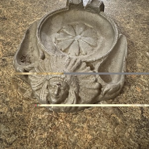 Puede incluir: Un cenicero gris de hierro fundido con un diseño floral en el centro y una cara nativa americana en el lateral. El cenicero tiene un cuenco circular y un borde decorativo.