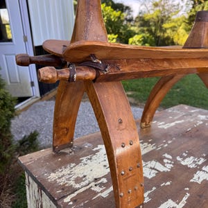 May include: A wooden saddle frame featuring curved legs, horizontal side rails, and two vertical pommel-like posts. The structure is held together with metal rivets, leather straps, and dark metal caps on the top of each post.
