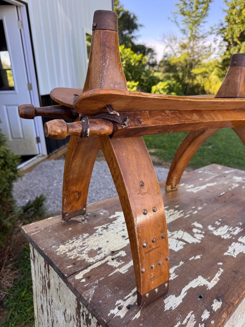 May include: A wooden saddle frame featuring curved legs, horizontal side rails, and two vertical pommel-like posts. The structure is held together with metal rivets, leather straps, and dark metal caps on the top of each post.