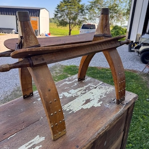 May include: A vintage wooden saddle-shaped stool featuring a curved seat, four arched legs reinforced with metal rivets, and two horizontal wooden dowels extending from the sides, all finished in a worn, natural brown wood tone.