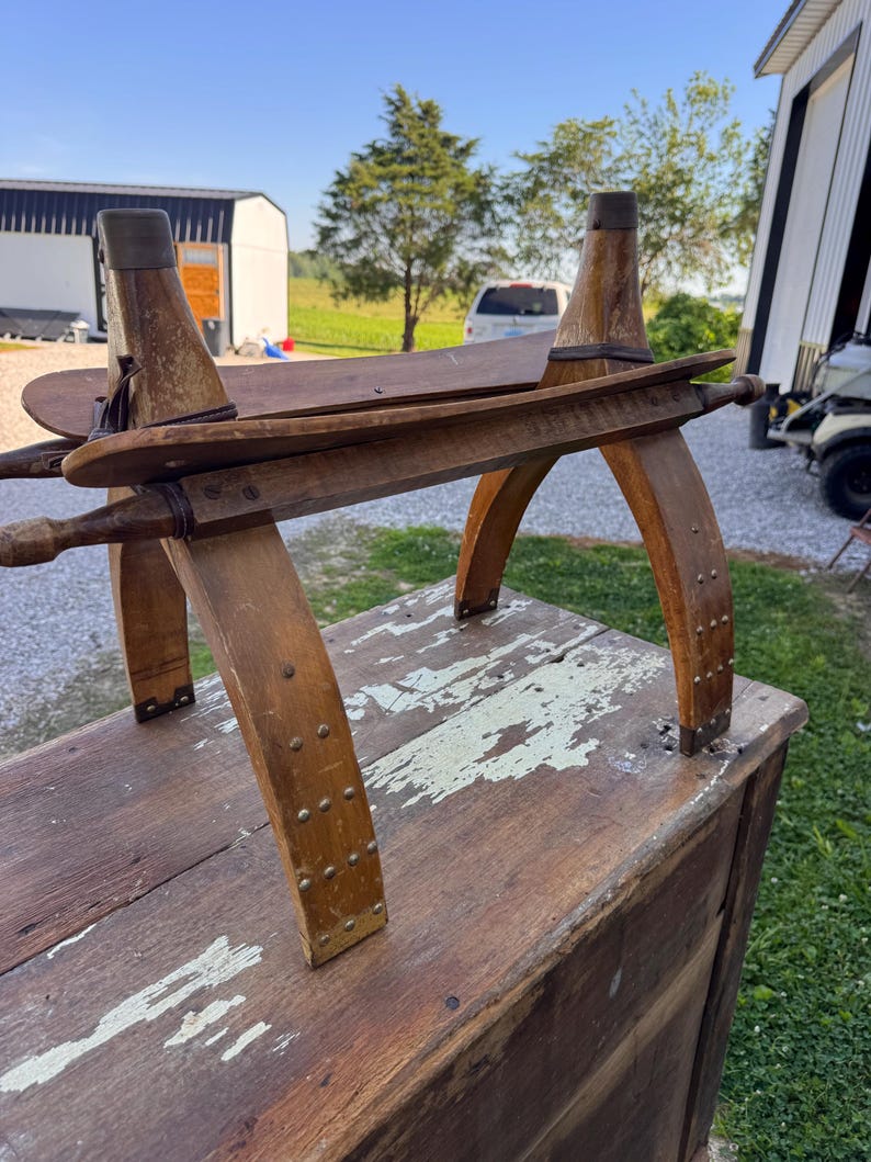May include: A vintage wooden saddle-shaped stool featuring a curved seat, four arched legs reinforced with metal rivets, and two horizontal wooden dowels extending from the sides, all finished in a worn, natural brown wood tone.