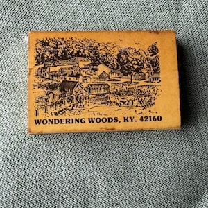 May include: A rectangular matchbox with a tan surface featuring a black ink illustration of a rural village scene with a water wheel and trees. The text WONDERING WOODS, KY. 42160 is printed in black capital letters at the bottom of the matchbox.