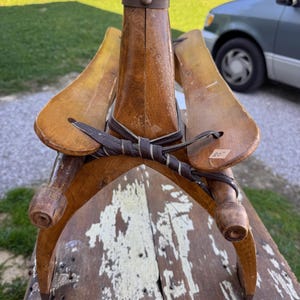 May include: A vintage wooden boot tree or shoe stretcher featuring a central vertical post with a dark brown cap, two curved side panels, and a dark brown leather strap tied in a knot across the front. A small white price tag marked 20 is attached.