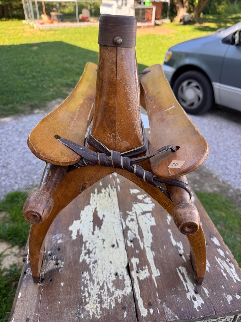 May include: A vintage wooden boot tree or shoe stretcher featuring a central vertical post with a dark brown cap, two curved side panels, and a dark brown leather strap tied in a knot across the front. A small white price tag marked 20 is attached.