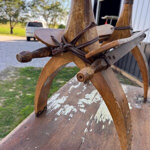 May include: A vintage wooden pack saddle featuring curved side bars, vertical wooden posts with metal caps, and dark brown leather straps secured in a cross pattern. The wood shows signs of wear with visible scratches and aged grain texture.
