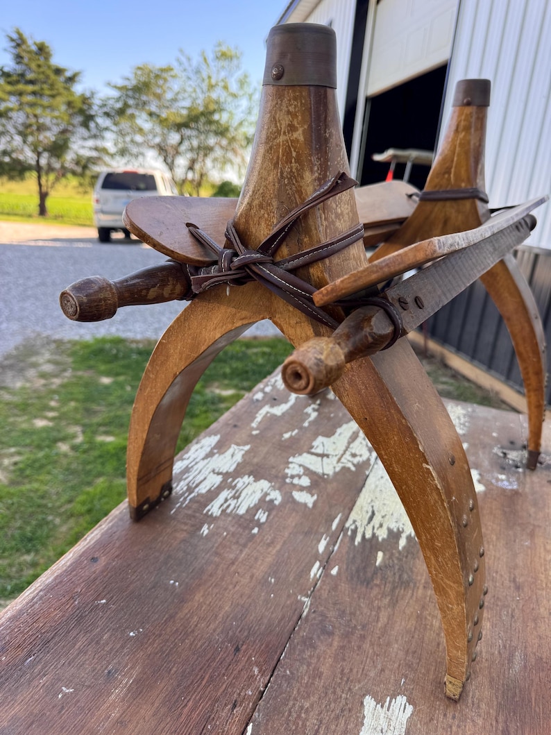 May include: A vintage wooden pack saddle featuring curved side bars, vertical wooden posts with metal caps, and dark brown leather straps secured in a cross pattern. The wood shows signs of wear with visible scratches and aged grain texture.
