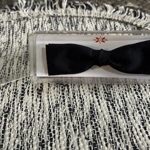 Puede incluir: Una pajarita negra en una caja de plástico transparente. La pajarita está centrada en una tarjeta blanca. La caja tiene un diseño floral rojo y dorado. La pajarita está sobre una superficie texturizada en blanco y negro.