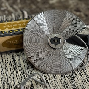 May include: A vintage Honeywell Tilt-A-Mite flash unit with a silver, fan-shaped reflector. The unit is accompanied by its original box, which has a black and gold color scheme. The flash unit is connected to a cord and is placed on a patterned surface.