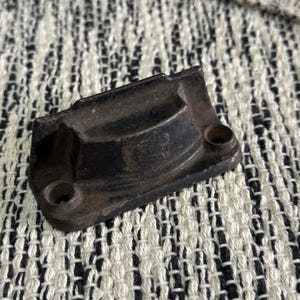 May include: A weathered, dark-colored metal object with a curved top and two holes. The object appears to be a small, rectangular component, possibly a part of a larger mechanism or device. The surface shows signs of wear and age.