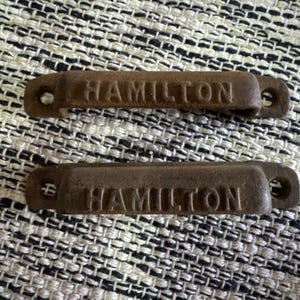 May include: Two antique, rectangular metal drawer pulls with the name "HAMILTON" embossed on each. The pulls are a weathered, rusty brown color, with holes on each end for mounting. They rest on a textured, neutral-toned fabric.
