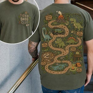May include: Olive green t-shirt with a dinosaur map design on the back and "Tired Dads Club" on the front. The map features dinosaurs, a volcano, and a jeep. The shirt is made of a soft material.