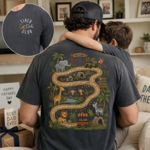 May include: A charcoal gray t-shirt featuring a small front chest graphic that reads TIRED DADS CLUB with a palm tree. The back displays a large, colorful jungle scene with a winding road, an elephant, giraffe, lion, zebra, monkeys, and a welcome sign.