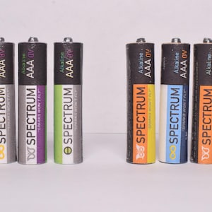 May include: A collection of AAA alkaline batteries with the brand name "SPECTRUM" printed on them. The batteries have a black top and a colorful design with the text "SPARKS WON'T FLY". The batteries are in various colors, including purple, green, and orange.