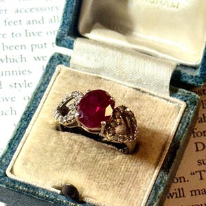 May include: A vintage ring featuring a large, oval ruby gemstone at its centre, complemented by smaller diamonds, set in a gold band. The ring is presented in a blue velvet jewellery box with a cream interior, highlighting its elegance.